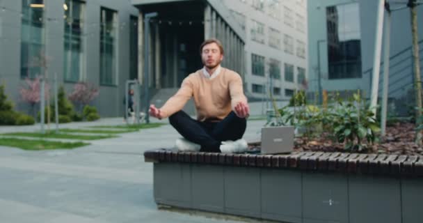 Bonito homem barbudo caucasiano de óculos relaxantes, sentado na posição de lótus no banco do pátio do prédio de escritórios ou da universidade. Estilo de vida saudável, ao ar livre — Vídeo de Stock