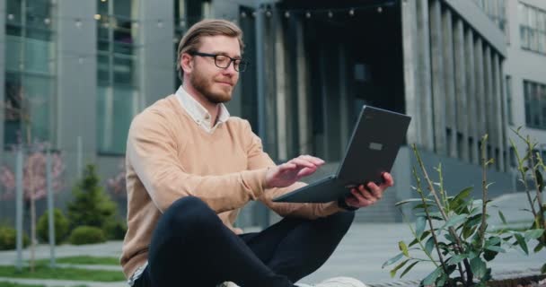 Bonito jovem caucasiano do sexo masculino trabalhando ou estudando usando laptop enquanto sentado cruzando pernas no banco do distrito financeiro da cidade. Ao ar livre — Vídeo de Stock