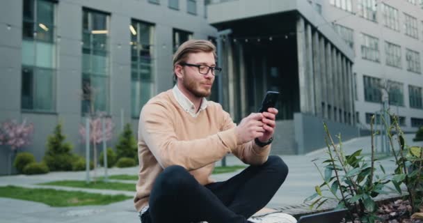 Framgångsrik kaukasier man i glasögon med mobiltelefon medan du sitter korsar ben på bänken i stadens finansdistrikt. Utomhus, man skriva online sms på telefonen sitter på en bänk — Stockvideo