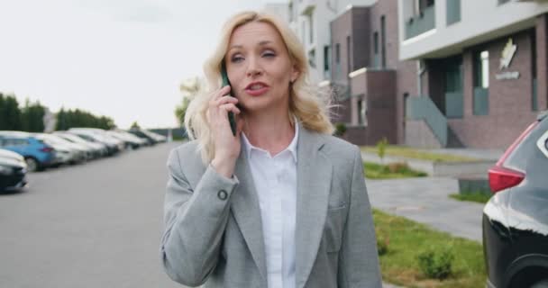 白人の自信に満ちたビジネスマンの女性が街を歩き、屋外のオフィスに行く携帯電話で話しています。ストリートでスマートフォンを使用してブロンドの中年女性. — ストック動画