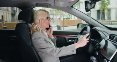 Şık giyinmiş neşeli iş kadını lüks bir arabada direksiyon başında akıllı telefondan konuşuyor. Kadın sürücü cep telefonu kullanıyor. Yavaş çekim.