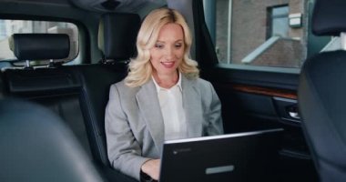 Modern arabada dizüstü bilgisayarda çalışan başarılı bir iş kadını. Beyaz, sarı saçlı, resmi takım elbiseli bayan arka koltukta oturuyor ve laptopta bilgi arıyor. Yavaş çekim.
