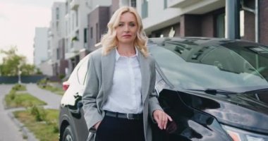 Modern binanın yanındaki otoparkta siyah arabanın yanında duran resmi giysiler içindeki başarılı beyaz iş kadınının portresi. Açık havada. Yavaş çekim
