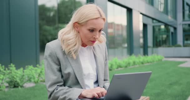 ブロンドの中年女性の勝者はラップトップを見てオンライン成功を祝うオフィスビルの屋外の背景に座っています ビジネスマンはノートパソコンでいい知らせを読んでいる スローモーション — ストック動画