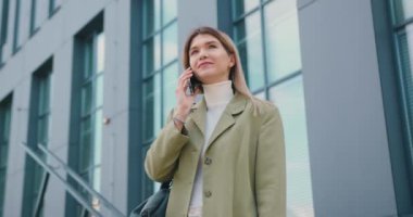 Gülümseyen genç bir kadın modern cam ofis binasının önünde duruyor ve cep telefonuyla video görüşmesi yapıyor. Kız gülüyor ve iyi haberlerden hoşlanıyor, dışarıda arkadaşlarıyla iletişim kuruyor. Yavaş çekim.