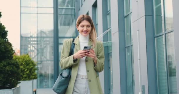 在下班后使用手机短信写作的同时 微笑专业的女商人 女孩使用智能手机打字短信走路背景现代建筑 慢动作 — 图库视频影像
