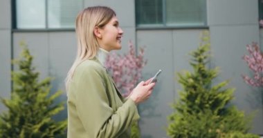 Dışarıda cep telefonu uygulaması kullanan mutlu genç bir kadının portresi. Yürüyen ve mesaj atan bir bayan yönetici ve ofis binasının yakınındaki haber yayını. Yavaş çekim.