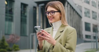 Şehir caddesinde akıllı telefon kullanarak şık günlük kıyafetler giyen güzel bir iş kadını portresi. Dışarıda elinde cep telefonu tutan çekici bir kız. Yavaş çekim.