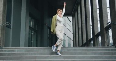 Ceket tutan güzel, gülümseyen kız merdivenlerden çıkıp dans ederek eğleniyor. Neşeli yaşam tarzı konsepti. Yavaş çekim.