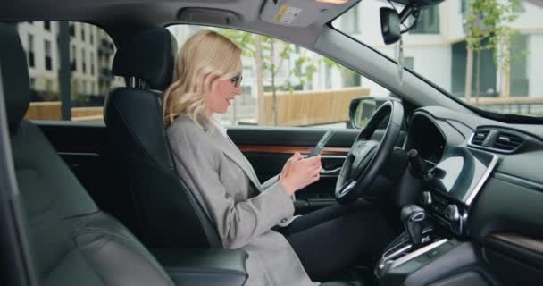 坐在司机座位上边开车边用智能手机打字边笑着的成年女人 集中精神的女商人在车里用手机的侧视图 慢动作 — 图库视频影像