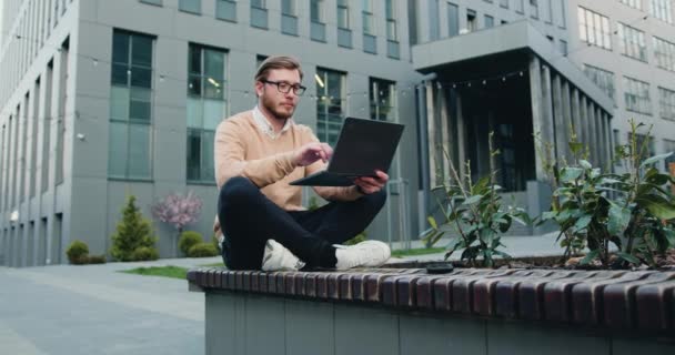 オフィスビルの近くの屋外に座ってノートパソコンを使用して若い男の背景 テクノロジーと都市生活の概念 スローモーション — ストック動画