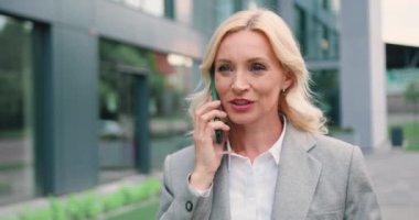 Kendine güvenen ve gülümseyen beyaz kadın portresi. Resmi giysiler içinde sokakta yürüyor ve cep telefonuyla konuşuyor. Yavaş çekim