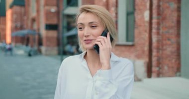 Kapalı kapılar ardında gülümseyen genç ve güzel bir iş kadını sokakta cep telefonuyla konuşuyor. Açık havada akıllı telefon kullanan neşeli sarışın kadın. Yavaş çekim.