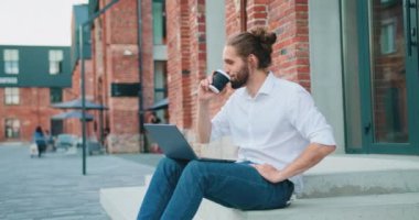 Rahat iş adamı, beyaz gömlekli bir andrepreneur, dışarıda çalışıyor, merdivenlerde oturuyor, kahve içiyor ve dizüstü bilgisayarda yazıyor. İş teknolojisi eğitim kavramı. Yavaş çekim.