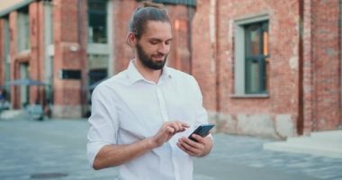 Yakışıklı, gülümseyen sakallı bir adamın portresi. Şehir caddesinde, açık havada, mesafeye bakarken cep telefonuyla yazı yazıyor. Yavaş çekim