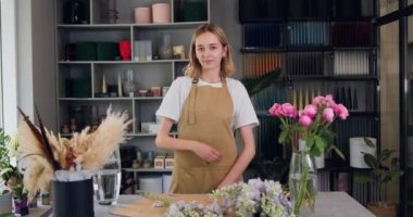 Portrait of a young woman business owner, smiling and looking at camera in a greenhouse. Gardener working in flower shop, plant store. Slow motion.