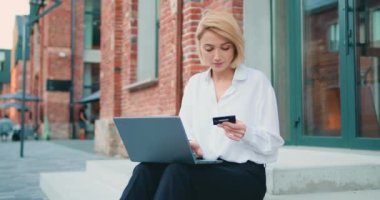 Bilgisayarı ve kredi kartıyla internetten alışveriş yapan genç bir kadın ofis binasının merdivenlerinde oturuyor. Laptop ve kredi kartıyla çalışan kendine güvenen güzel bir kadın, serbest çalışan. Yavaş