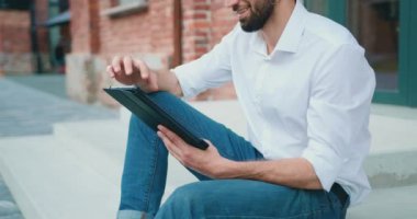 Smiling young man in casual wear use modern touch pad device use wireless internet while sitting on stairs at building background.Positive male employee in reading news on digital tablet. Outdoors