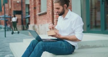 Young handsome guy eating takeaway healthy salad outdoors and watching videos on laptop, relaxing on stairs in front of office building on street. Slow motion