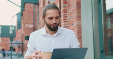 Portrait of attractive business man eating takeaway healthy food outdoors and using laptop, standing in front of office building on street. Slow motion