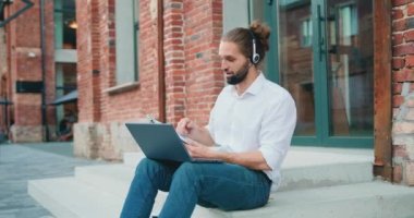 Attractive male manager in headset leading working meeting through video call on laptop while sitting on stairs outdoor. Slow motion