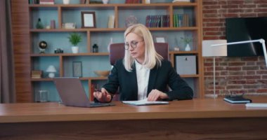 Portrait of good-looking purposeful qualified business lady in fromal wear which sitting at the table in well-equipped workplace and writing down datas from computer into financial documents