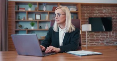 Beautiful experienced purposeful female office manager in black jacket sitting at her workplace in well-equipped cabinet and typing on computer,later thinking about something looking away