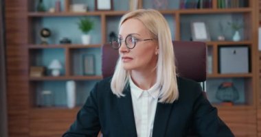 Adorable smiling skilled adult female office manager in formal business wear first look into window and later posing on camera in modern well-equipped cabinet,business portrait
