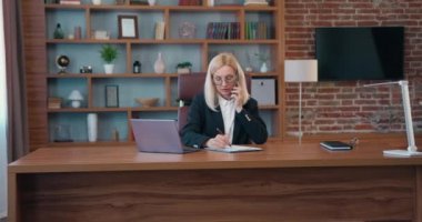 Kendine güveni tam sarışın iş kadını portresi. İyi donanımlı çalışma alanında masada oturan ve bilgisayarla aynı anda akıllı telefondan konuşan.