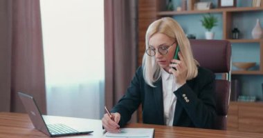 İş konsepti, resmi giyinmiş gözlüklü, deneyimli yetişkin bir iş kadınının bilgisayardaki çalışmalarını finansal raporlar ve mobil konuşmalarla birleştirmesi.