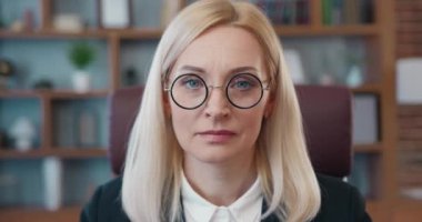 İyi donanımlı bir ofiste gözlüğünü çıkarırken kameraya bakan başarılı yetişkin kadın ofis müdürünün iş portresi.