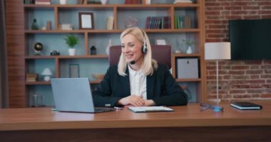 Pretty positive high-skilled effective blond female office manager wears headphones with mic sitting at her workplace in well-equipped office during online meeting with business partners using