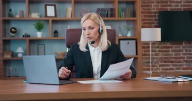 Workflow in office room where attractive experienced purposeful skilled businesswoman using headphones with mic during online briefing on computer together with business partners and they