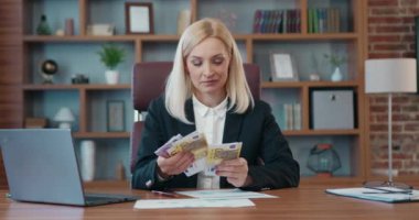 Portrait of charming happy successful light haired female ceo which enjoying calculating two hundred and five hundred euro banknotes,her business profit as a result of effective work