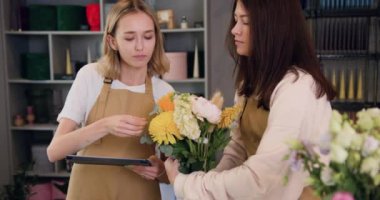 Women florists colleagues standing in flower shop with flowers, using digital tablet and make a bouquet. Small business concept. Slow motion.