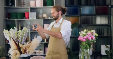Man florist using smartphone and taking photo of flowers. Caucasian bearded male florist holding potted flower in a modern flower shop. Slow motion.