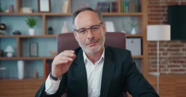 Portrait of good-looking positive high-skilled successful bearded ceo in business suit which taking off his eyeglasses and looking into camera with sincerely smile on office room background