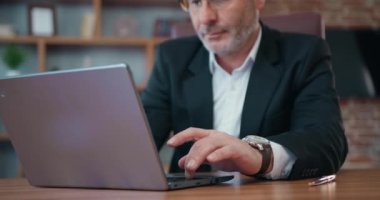 Good-looking confident responsible successful bearded 50s main office manager in eyeglasses wears business suit working on computer on modern office background,close view