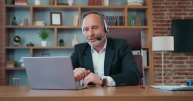Workflow in office where handsome motivated skilled purposeful 50s bearded office manager in formal wear using headphones with mic talking with colleagues via videocall on laptop