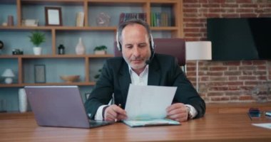 Good-looking responsible skilled successful 50s bearded head of company in headphones with mic holding video meeting with coworkers using computer and get acquainted with results of research