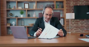 Handsome high-skilled hardworking successful 50s bearded businessman in headphones with mic holding video meeting on laptop with colleagues and explaining charts with datas