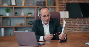 Business life concept where attractive skilled responsible 50s bearded main office manager in formal wear using headphones with mic talking with partners via videocall on computer and discussing