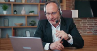 Business concept where handsome experienced responsible 50s bearded office manager in headphones with mic talking with colleagues using videochat on computer and summarizing results of briefing