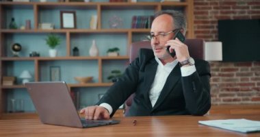 Good-looking experienced successful adult bearded company manager sitting at front of laptop,typing on it and talking on smartphone in well designed office,front view
