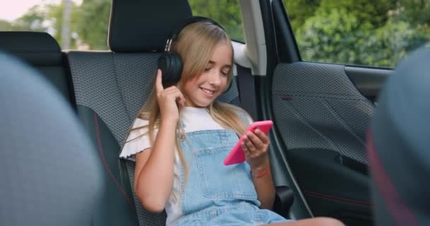 一个时尚的小女孩坐在车里 一边听音乐 一边用智能手机在后座上听音乐 慢动作 — 图库视频影像