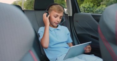 10 yaşındaki çocuk kulaklık takıyor, tablet kullanıyor, müzik dinliyor ve mutlu bir şekilde dans ediyor. Çocuk modern bir arabanın arka koltuğunda oturuyor ve dijital tablet kullanıyor. Yavaş çekim.