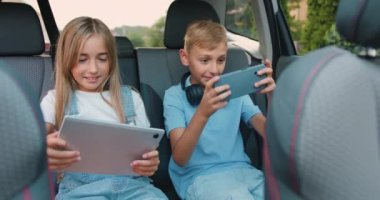Güzel kız ve erkek çocuklar modern bir arabada otururken telefon ve tabletle oyun oynayarak eğleniyorlar. Küçük kız ve erkek kardeş gülüyor ve mutlu bir şekilde kutlama yapıyorlar.