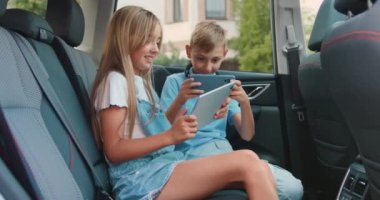 Attaractive oğlan ve kız, Car 'da tablet ve akıllı telefon oynuyor. Çocuklar oyun ve eğlence için bir cihaz kullanıyorlar, otomatikle seyahat ediyorlar. Yavaş çekim.