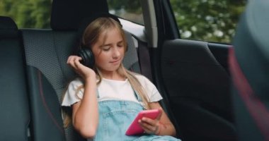 10 yaşında, kulaklıklı, gülümseyen ve cep telefonu kullanan bir kız. Arka koltukta otururken ve aile gezisi sırasında müzik dinlerken. Yavaş çekim.