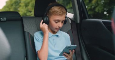 Çocuk arabada oturuyor ve akıllı telefon ve kulaklık kullanıyor. Aile gezisi sırasında arabanın arka koltuğunda oturmuş müzik dinleyen cici bir çocuk. Yavaş çekim.